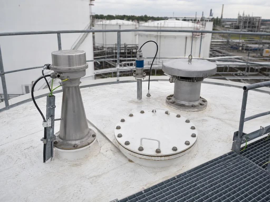 Fixed-roof storage tank platform showing a radar level transmitter, independent high-high level switch, gauge hatch, vent, conduit, and access handrails